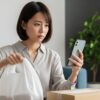 通販の荷物を前にスマートフォンでトコジラミの情報を調べている不安そうな日本人女性