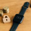 木製デスク上に置かれた充電切れのスマートウォッチとパドロックの置物、隣に積まれた硬貨が費用とセキュリティの不安を象徴する