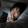 布団の中でスマートフォンをこっそり見ている20代の日本人女性。プリ小説を親バレせずに安心して楽しむという、記事のテーマを象徴する一枚。彼女の安心した表情が、安全対策を知ることの重要性を示唆している。
